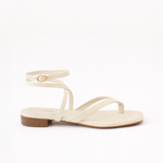 A single flat sandal in a light cream or off-white color, featuring a low block heel, a thong-style toe strap, and an ankle strap with a small gold buckle.