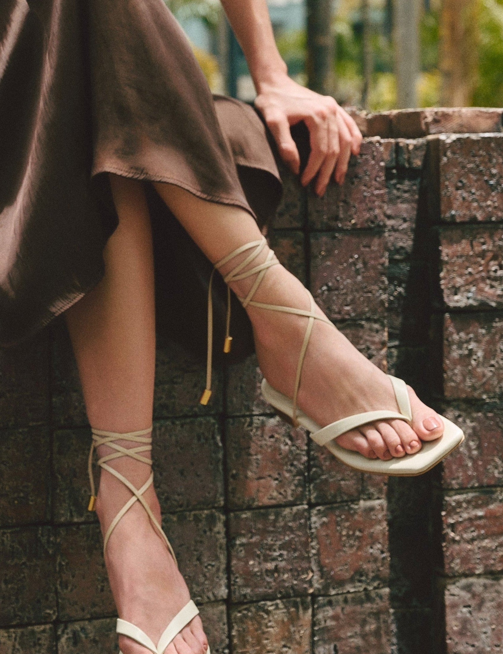 Person wearing a brown skirt and white cream leather sandals sitting on a stone wall.